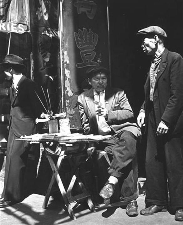 View of three Asian men in front of shop. The man in the center is seated behind a small folding table with newspapers and various items on top. - Circa 1940s